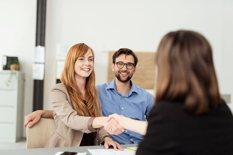 a happy woman shakes a financial counselor's hand as her husband sits behind her.